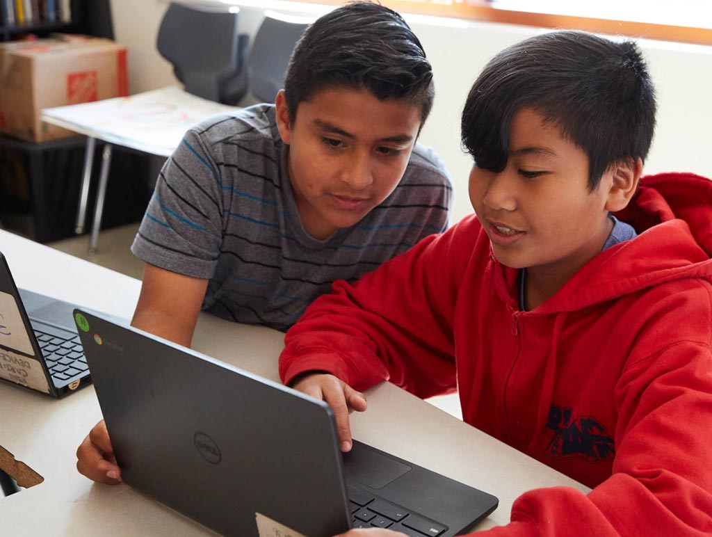 A Latino boy and Asian boy sit and look at a laptop together.