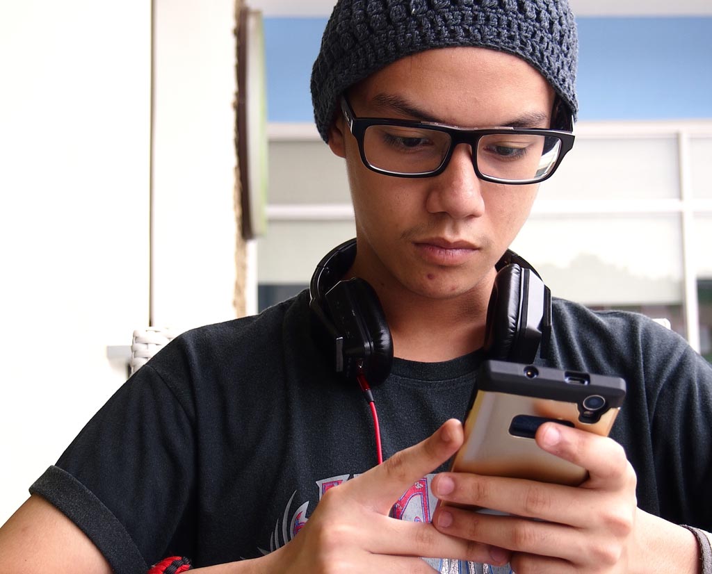 A teenage boy with glasses and beanie looks down at the cellphone he's holding, headphones draped around the back of his neck.