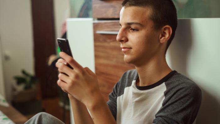 A teen boy sitting on the floor against a wall looking at his phone.