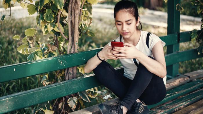 Teen girl using her smartphone while sitting on a park bench.