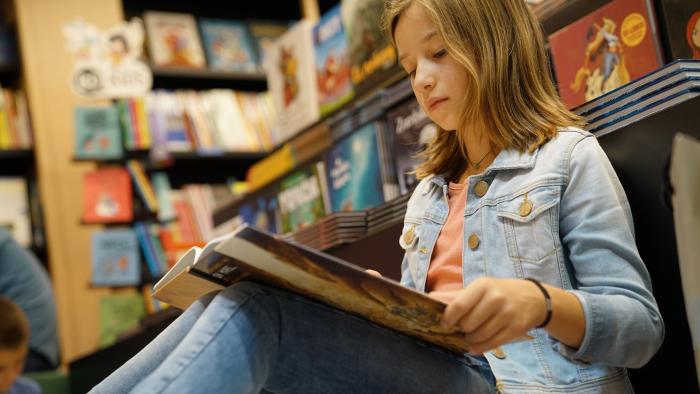 A young girl sitting on the floor of a bookstore with an open book in her lap.