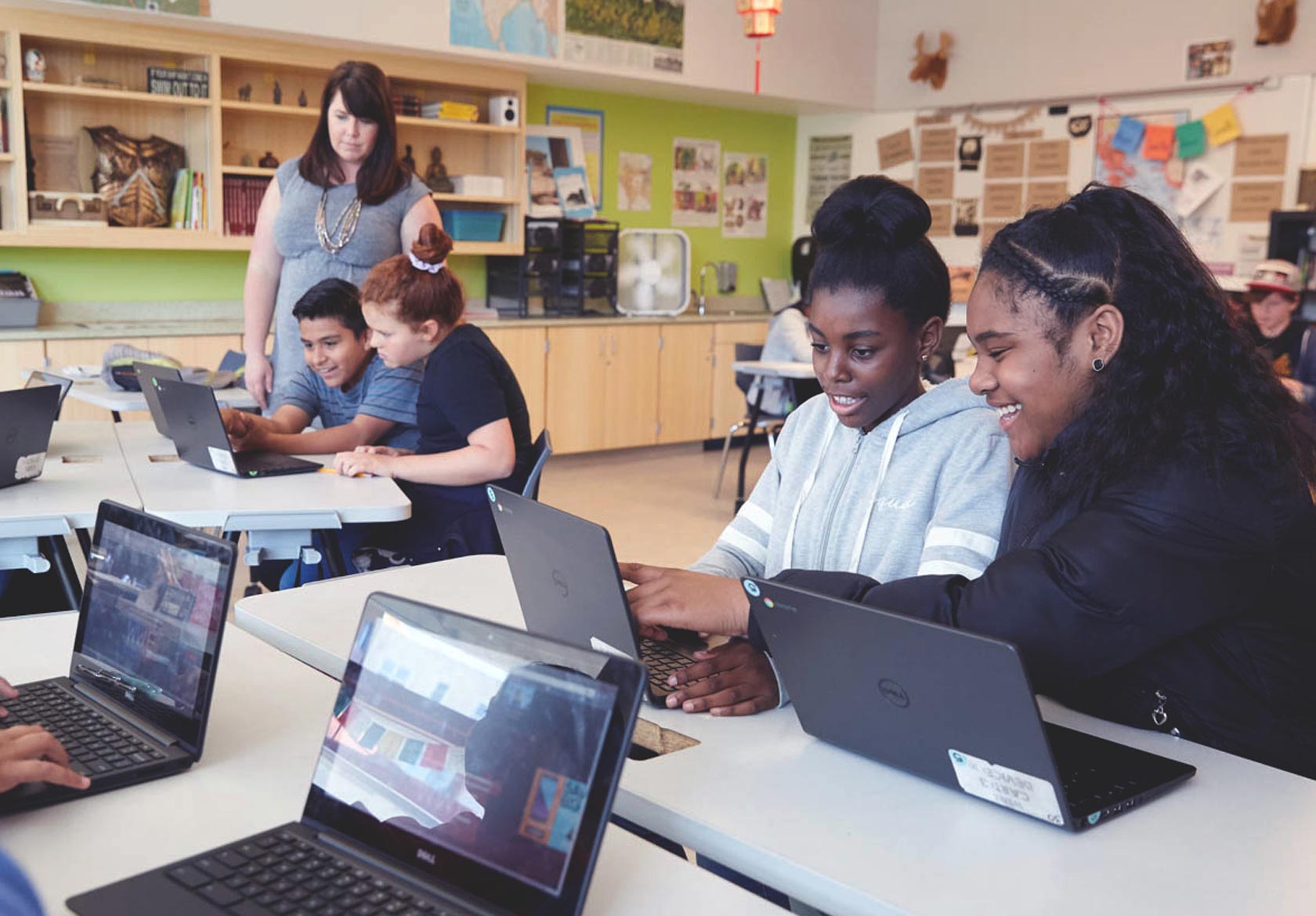 A classroom with two Black girls smiling and looking at a laptop, with a Latino boy, White girl, and female teacher in the background.