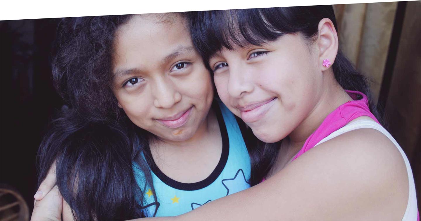 Two young teen girls with black hair and brown skin embrace and smile at the camera.