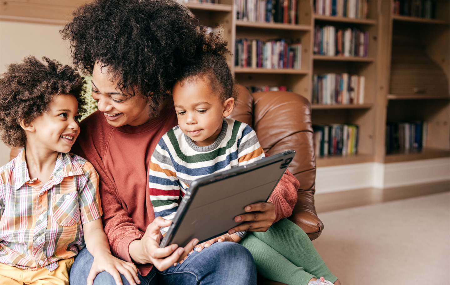 A Black woman smiles and holds a tablet as two Black children sit in her lap, bookcase in the background.