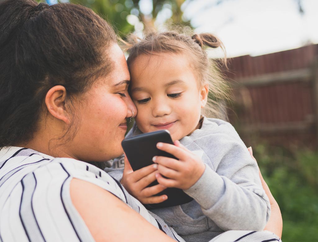 A Latina woman smiles with eyes closed, holding a girl as the girl looks at a cellphone.